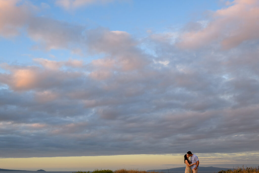 A couple embraces under a wide, colorful sky filled with soft clouds during a serene sunrise engagement on Maui